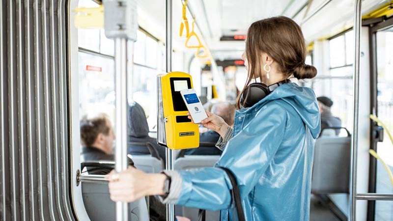Woman in a blue slicker tapping her mobile wallet at a fare reader on a bus to pay her public transit fare