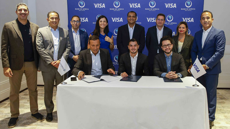 A group of eleven business professionals, smiling and posing. Two seated men sign papers at a table. Background shows "Bank of Africa" and "Visa" logos.